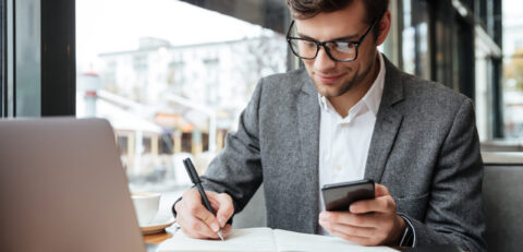 Smiling business man in eyeglasses sitting by the table in cafe with laptop computer while using smartphone and writing something